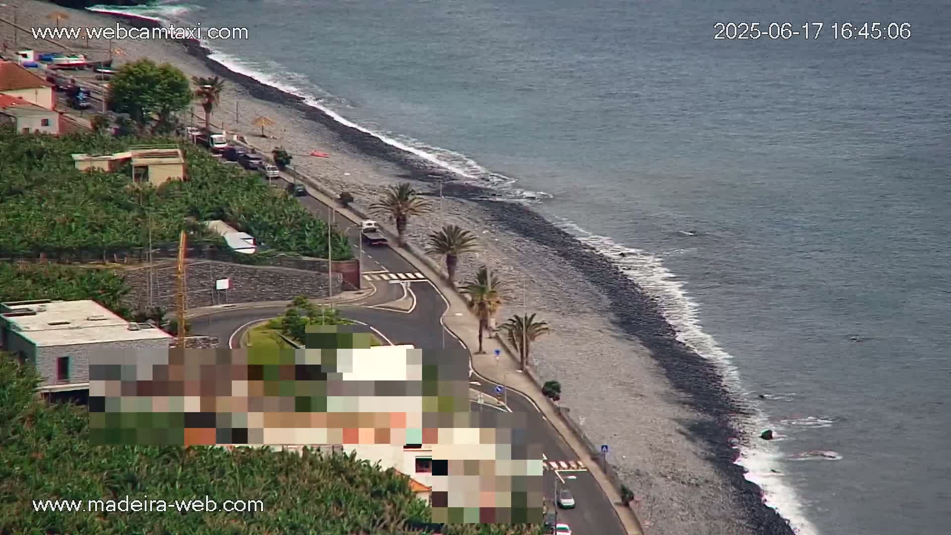 An aerial view shows a dark-grey pebble beach meeting a calm, grey-blue ocean, with a road and palm trees lining the shore, and buildings and vegetation inland.