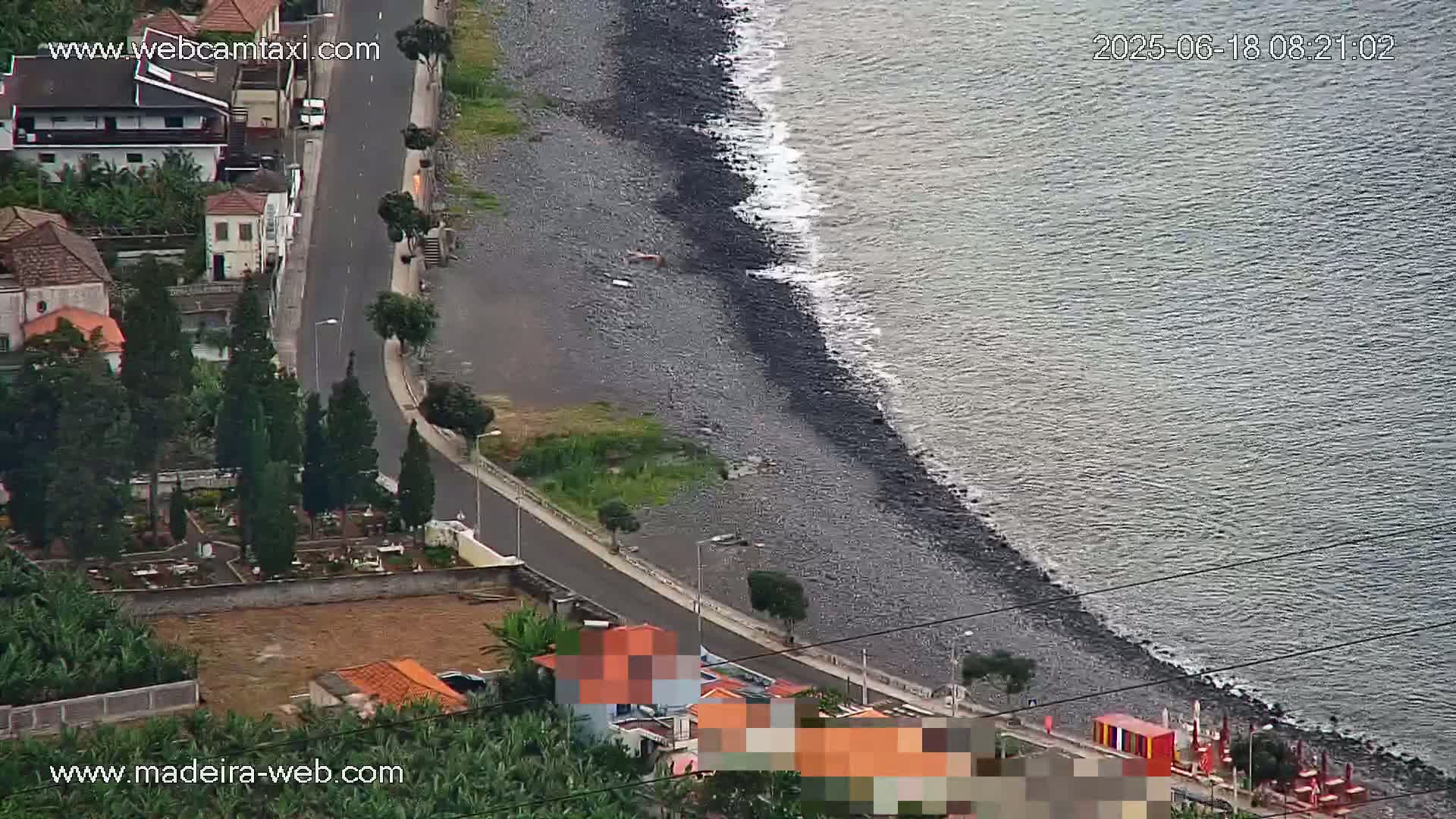 A dark-colored, rocky shoreline meets a calm ocean, next to a road with buildings and a cemetery visible above.
