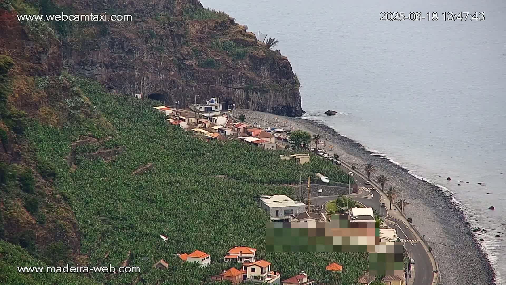 A coastal village nestled between a steep, rocky cliff and a dark-pebbled beach, with a calm ocean and banana plantations covering the hillside.