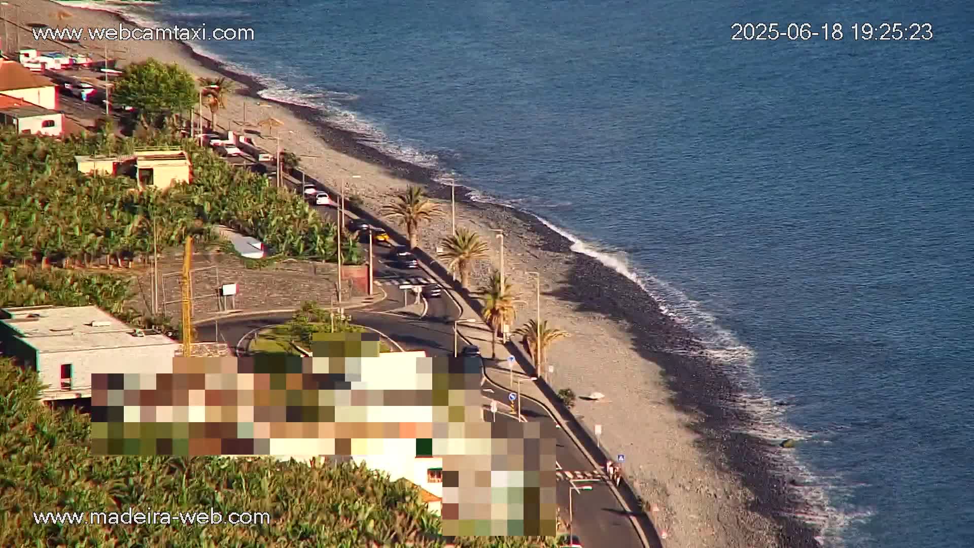An aerial view shows a dark-sand beach with gentle waves lapping the shore, a road curving alongside it, and palm trees lining the roadside next to buildings and vegetation.