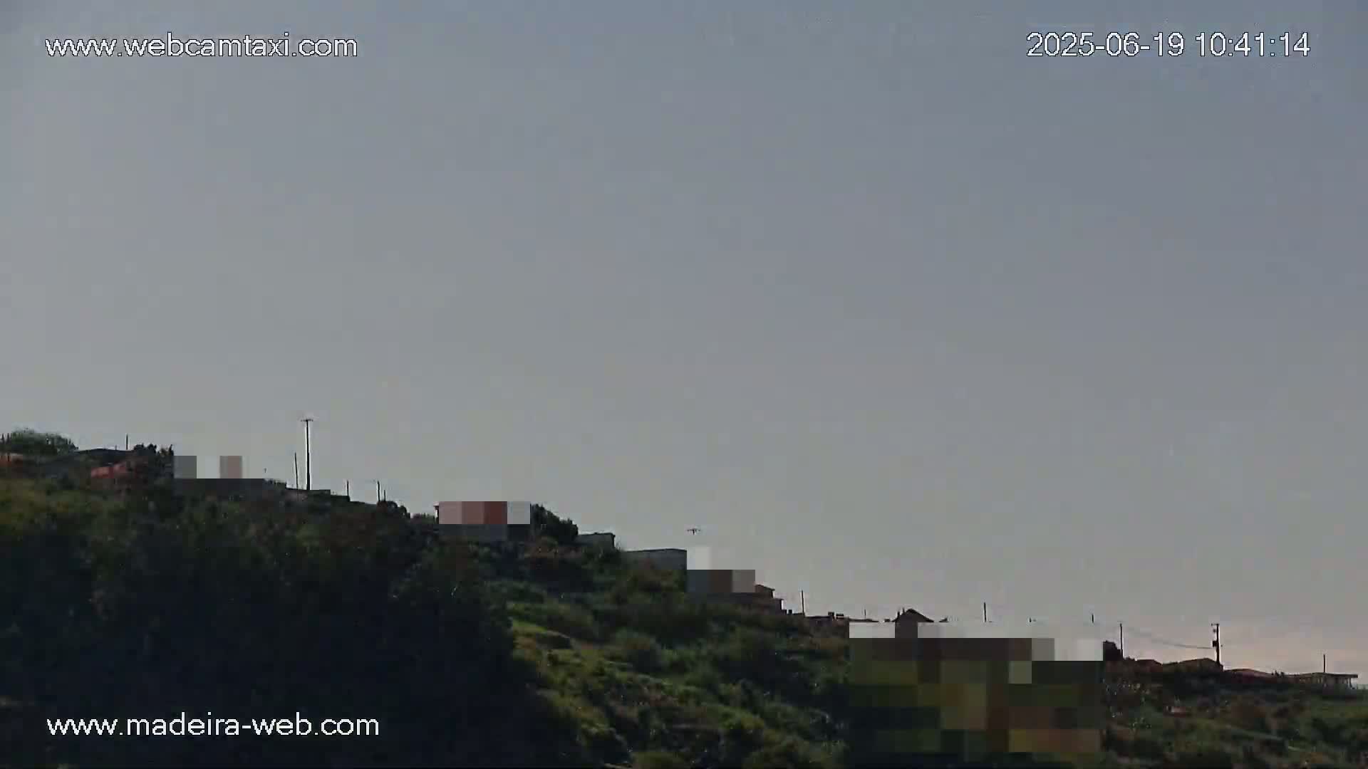 A distant airplane is visible in a hazy sky above a hillside with sparse buildings and dark green vegetation.