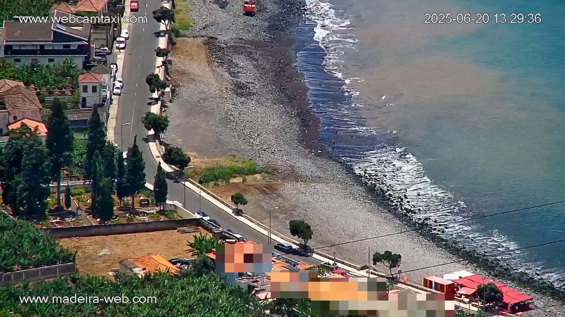 A rocky beach with gentle waves meets a road lined with buildings and trees on a sunny day.