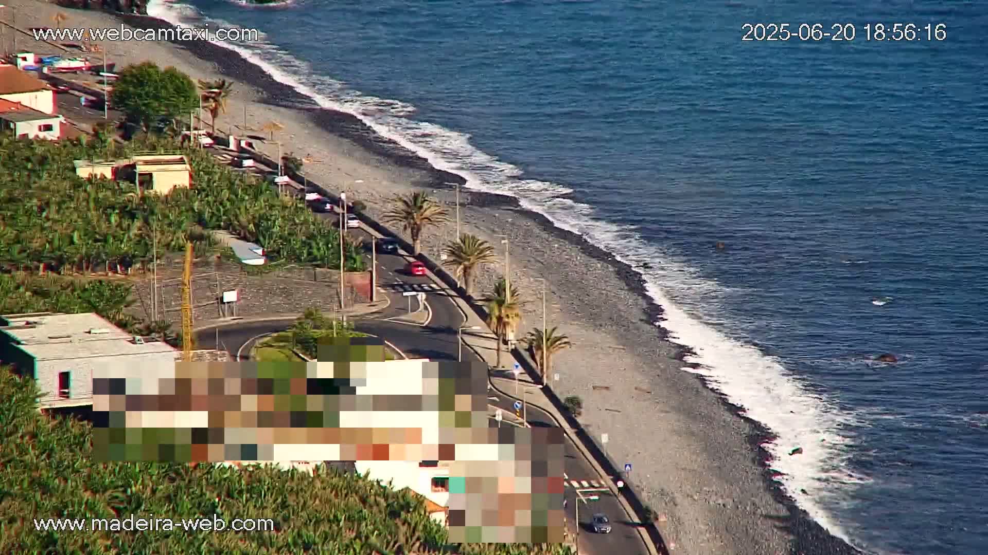 A coastal road curves along a pebble beach with gentle waves lapping the shore, palm trees lining the road and buildings visible beyond.