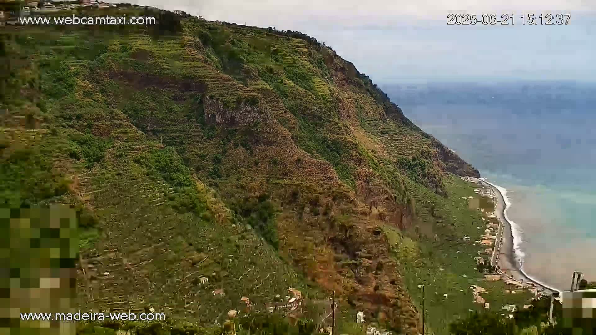 A verdant, terraced hillside descends to a dark-sand beach and ocean under a mostly clear sky.