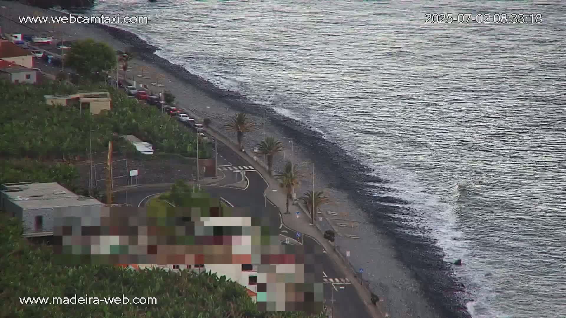 A dark-colored beach with moderate waves laps a shoreline road lined with palm trees and buildings, under a mostly clear sky.
