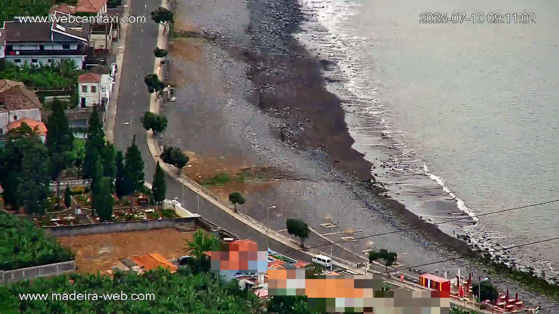 An aerial view shows a dark-colored, pebble beach meeting a calm, gray ocean next to a coastal road lined with trees and buildings, under an overcast sky.
