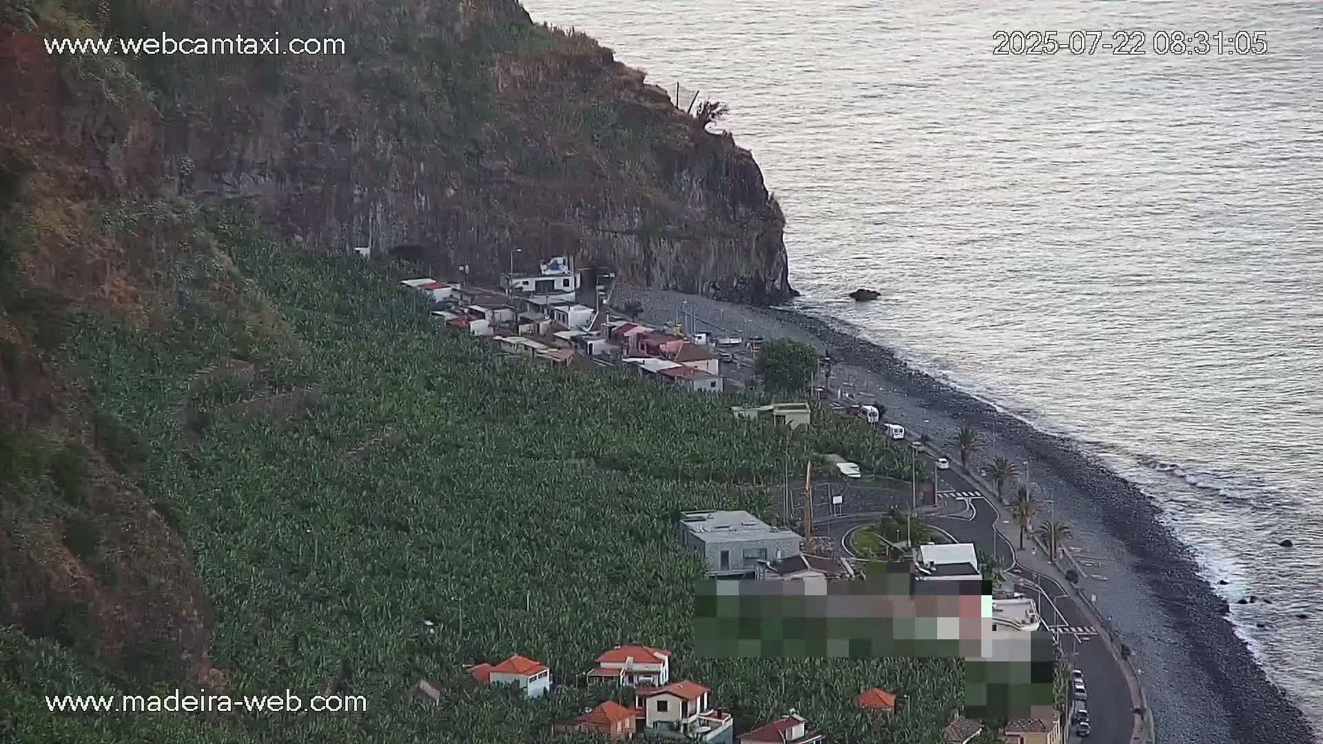 A coastal scene shows a rocky cliff overlooking a dark-sand beach with buildings and banana plantations on the hillside, under a calm, clear sky.