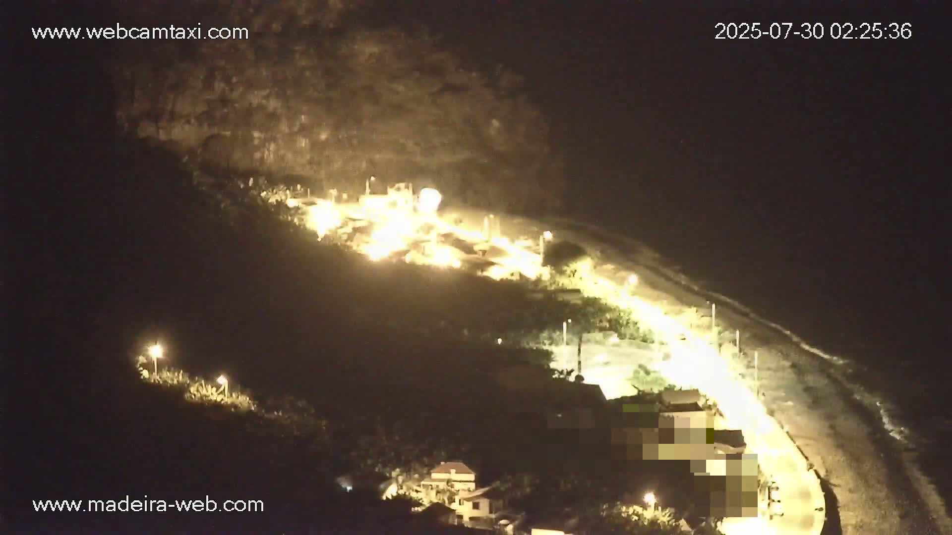 A coastal scene shows a rocky cliff overlooking a dark-sand beach with buildings and banana plantations on the hillside, under a calm, clear sky.