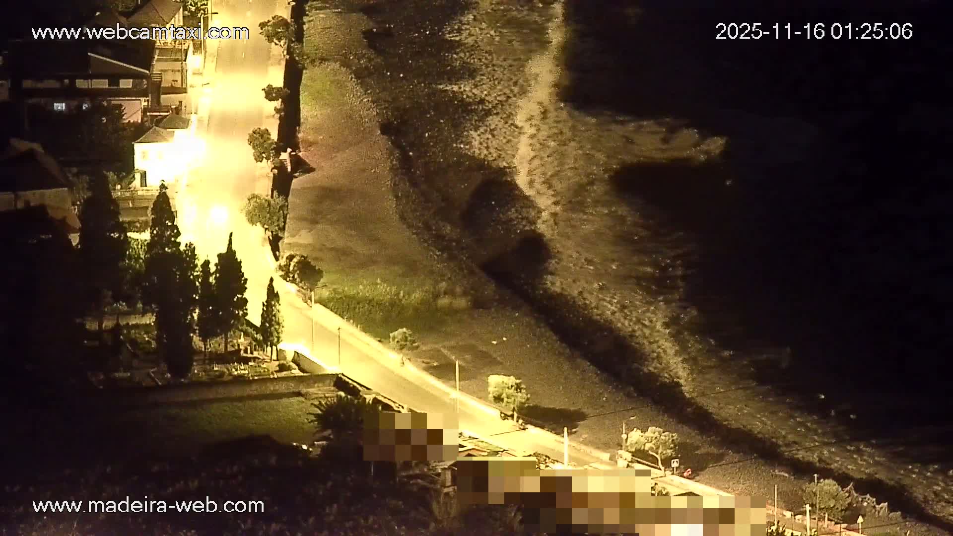 An overhead night view reveals an illuminated coastal road winding past buildings and trees, bordering a dark sea where faint waves break on the shore under clear skies.