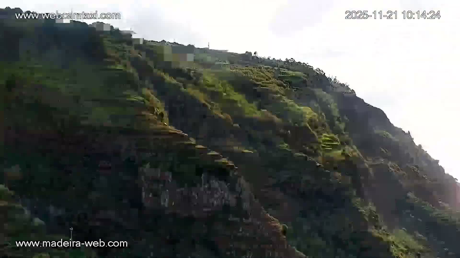A lush, terraced green mountainside with several buildings near its summit stretches under a bright, hazy, overcast sky.