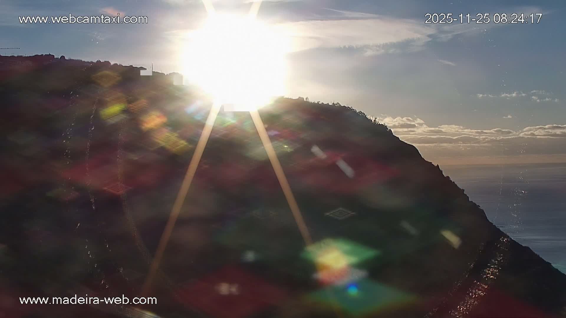 A brilliant sun low in the sky creates a strong sunburst and lens flare over a dark, steep coastal hillside with visible buildings and the calm ocean, under sunny, partly cloudy conditions.