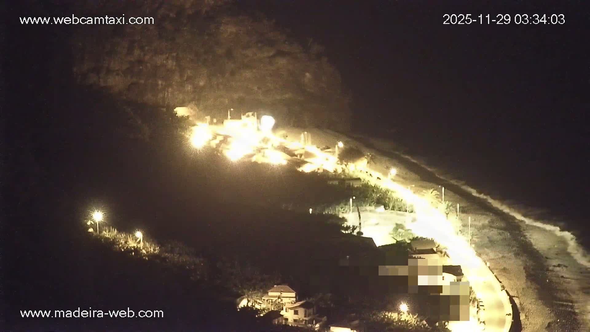 A brightly illuminated coastal settlement, featuring a prominent cluster of intense lights on a hillside, overlooks a curving road, numerous buildings, and waves breaking on the shore at night under clear skies.