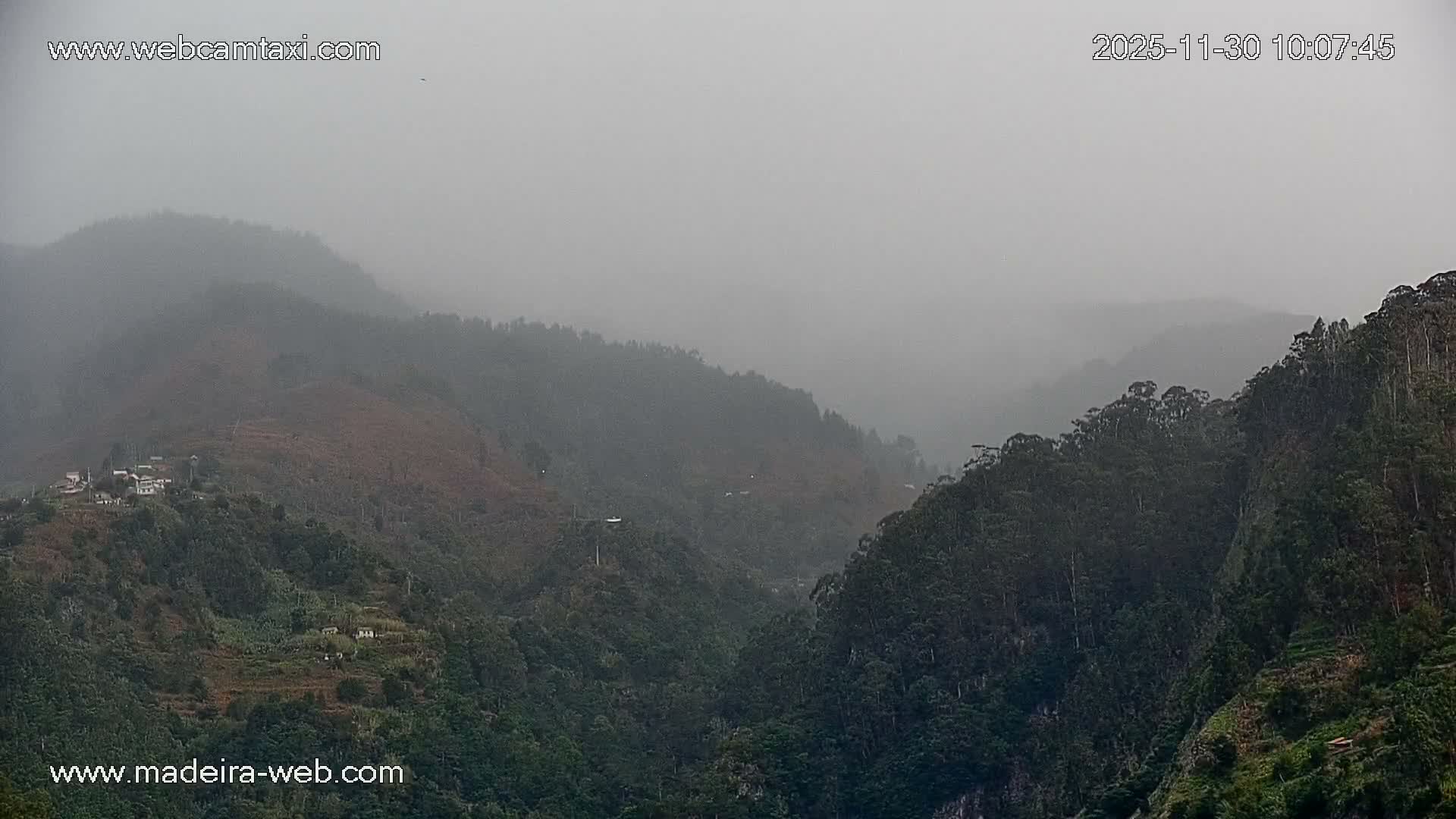 A vast, forested mountainous landscape is shrouded in mist under an overcast sky, with scattered small buildings visible on the lower slopes.