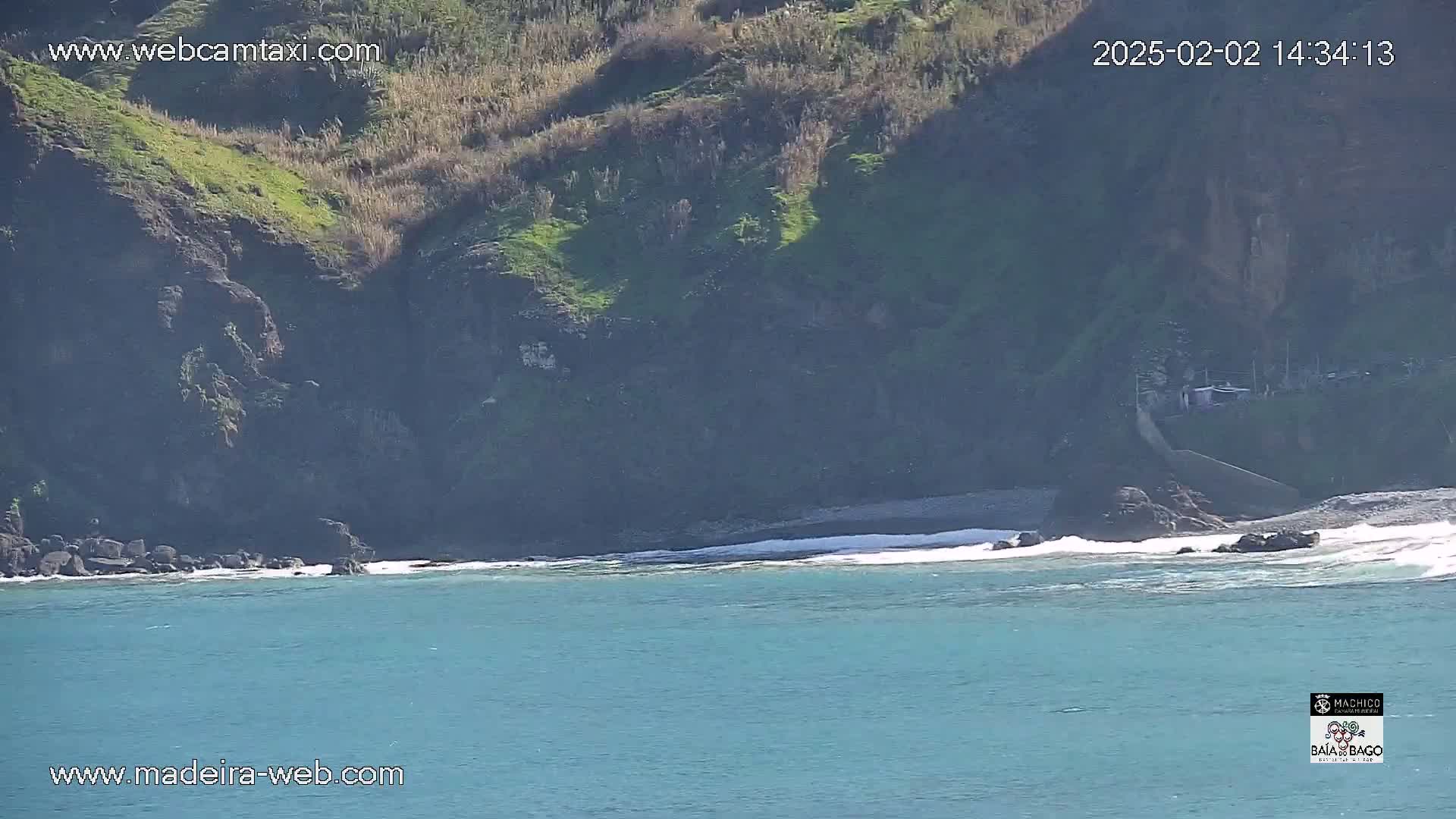 A rocky, green-covered cliff overlooking a calm, teal ocean with small waves lapping the shore on a sunny day.