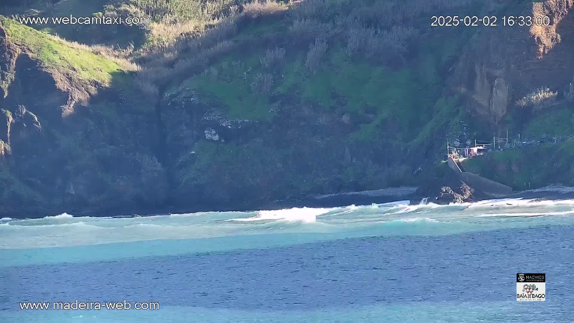A rocky, partially vegetated cliff overlooks a light-blue ocean with small waves breaking on a dark-colored beach under a bright, sunny sky.
