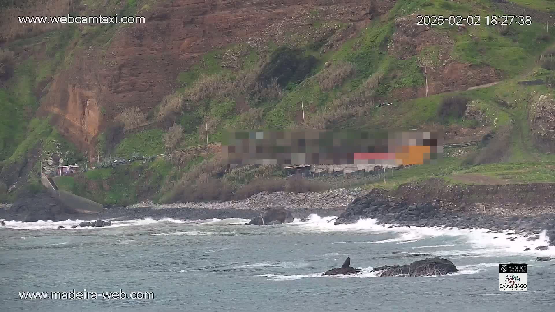 A rocky shoreline with crashing waves meets a steep, green hillside under overcast skies.