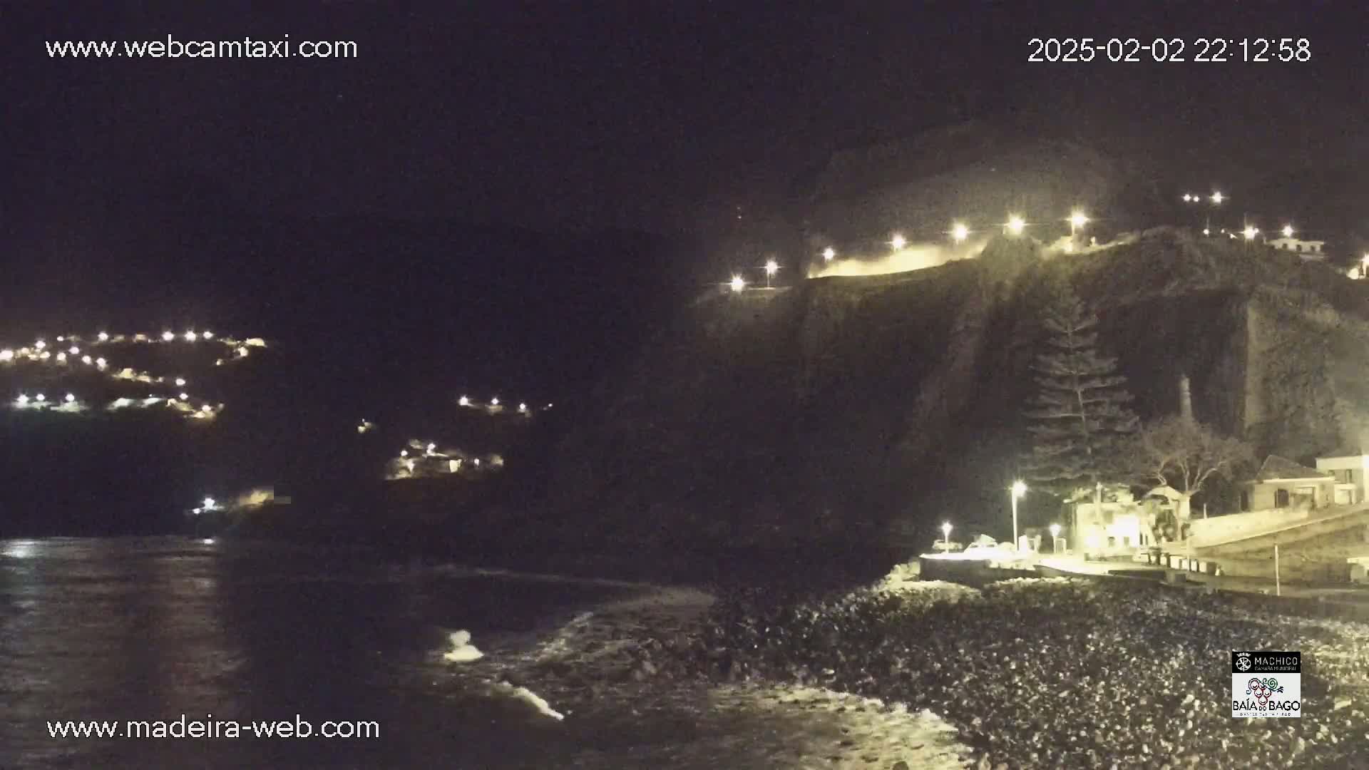 A nighttime coastal scene shows a dark cliffside with illuminated buildings and roads, overlooking a rocky shoreline with gentle waves under a clear, dark sky.