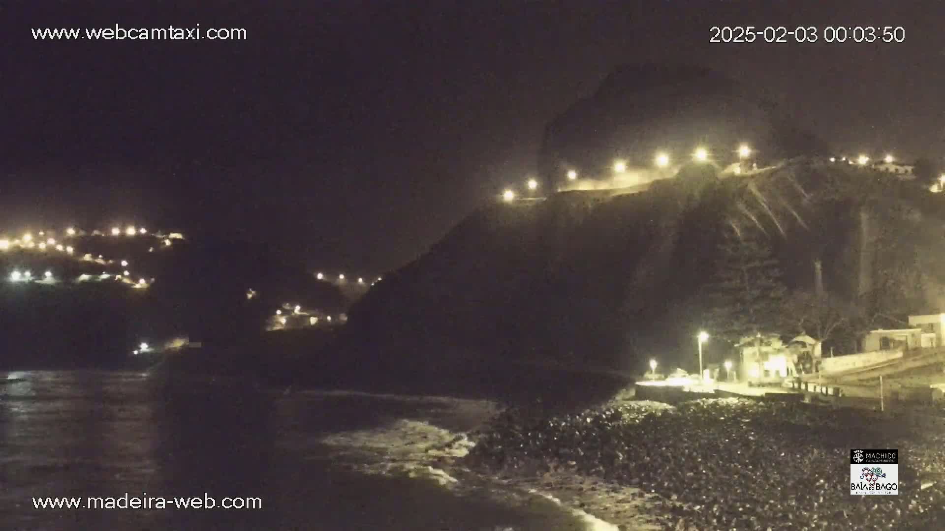 A nighttime view of a dark, rocky coastline with a village illuminated by lights on a hillside, gentle waves lapping at the shore.