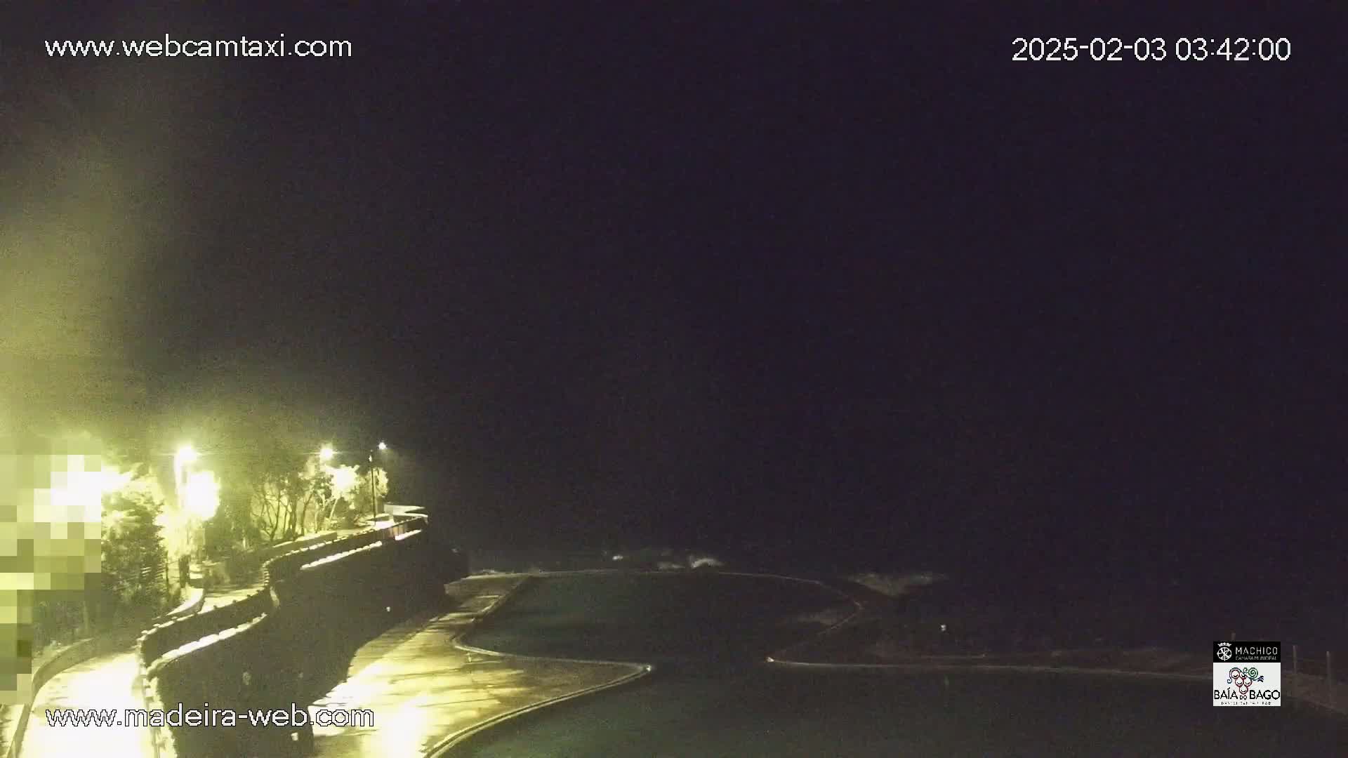A nighttime view of a dark ocean with waves crashing near a lit, empty outdoor pool and a paved walkway.
