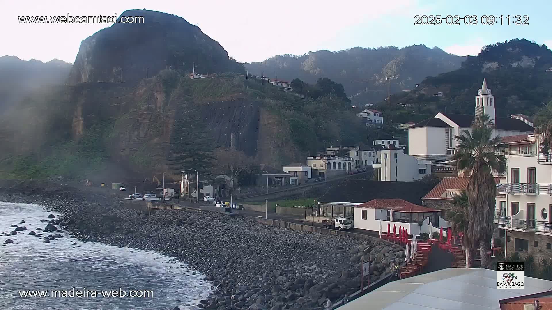 A rocky coastline with a village of white buildings nestled against a steep, dark cliffside under a hazy sky.