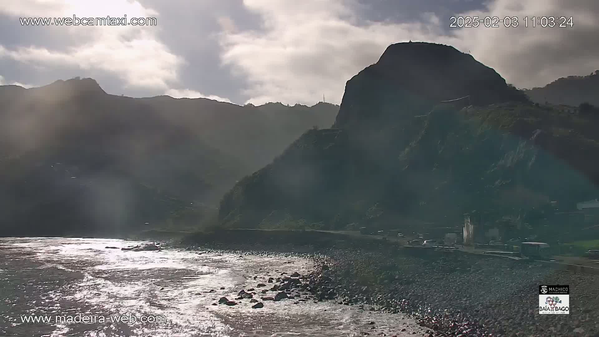 A rocky, dark coastline meets a slightly choppy ocean under a partly cloudy sky.