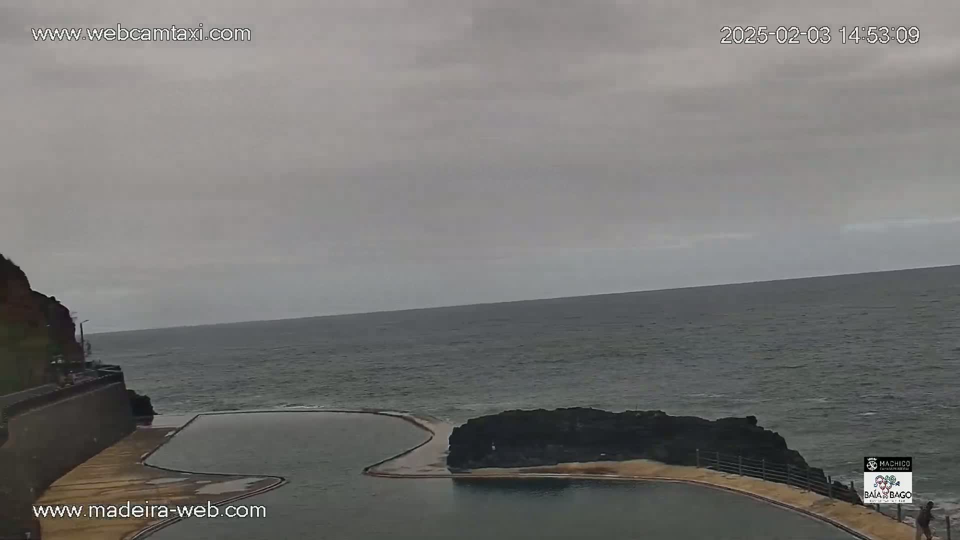 An oceanside swimming pool is situated on a rocky cliff overlooking a calm, grey sea under an overcast sky.
