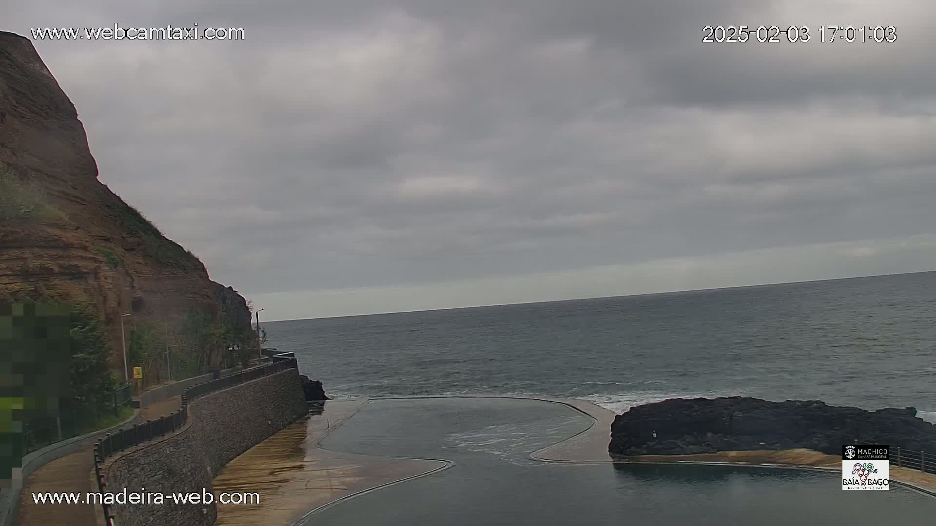 A rocky cliff overlooks a calm ocean with a dark-colored natural swimming pool nestled between the rocks, under an overcast sky.