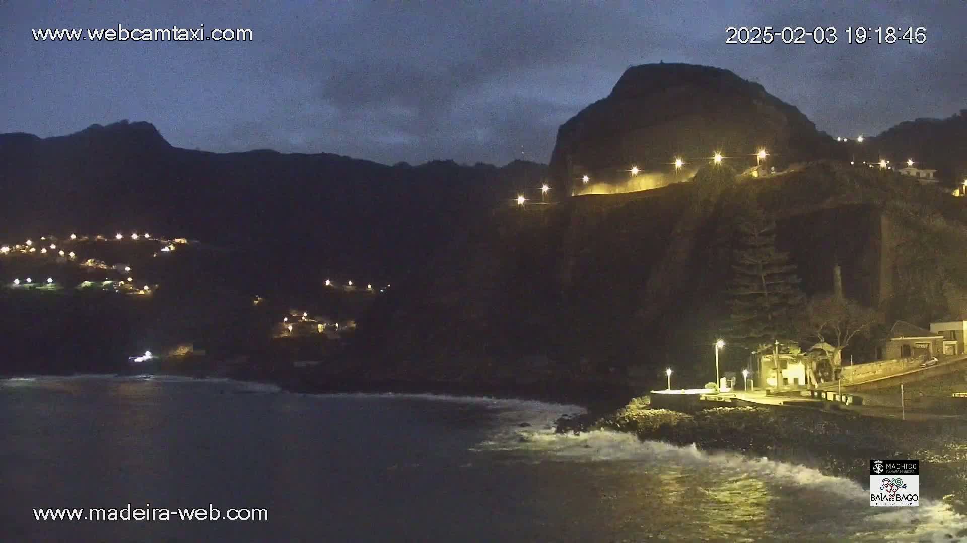 A dark, nighttime coastal scene shows a rocky cliffside with lights illuminating a road and buildings, while gentle waves lap the shore beneath.