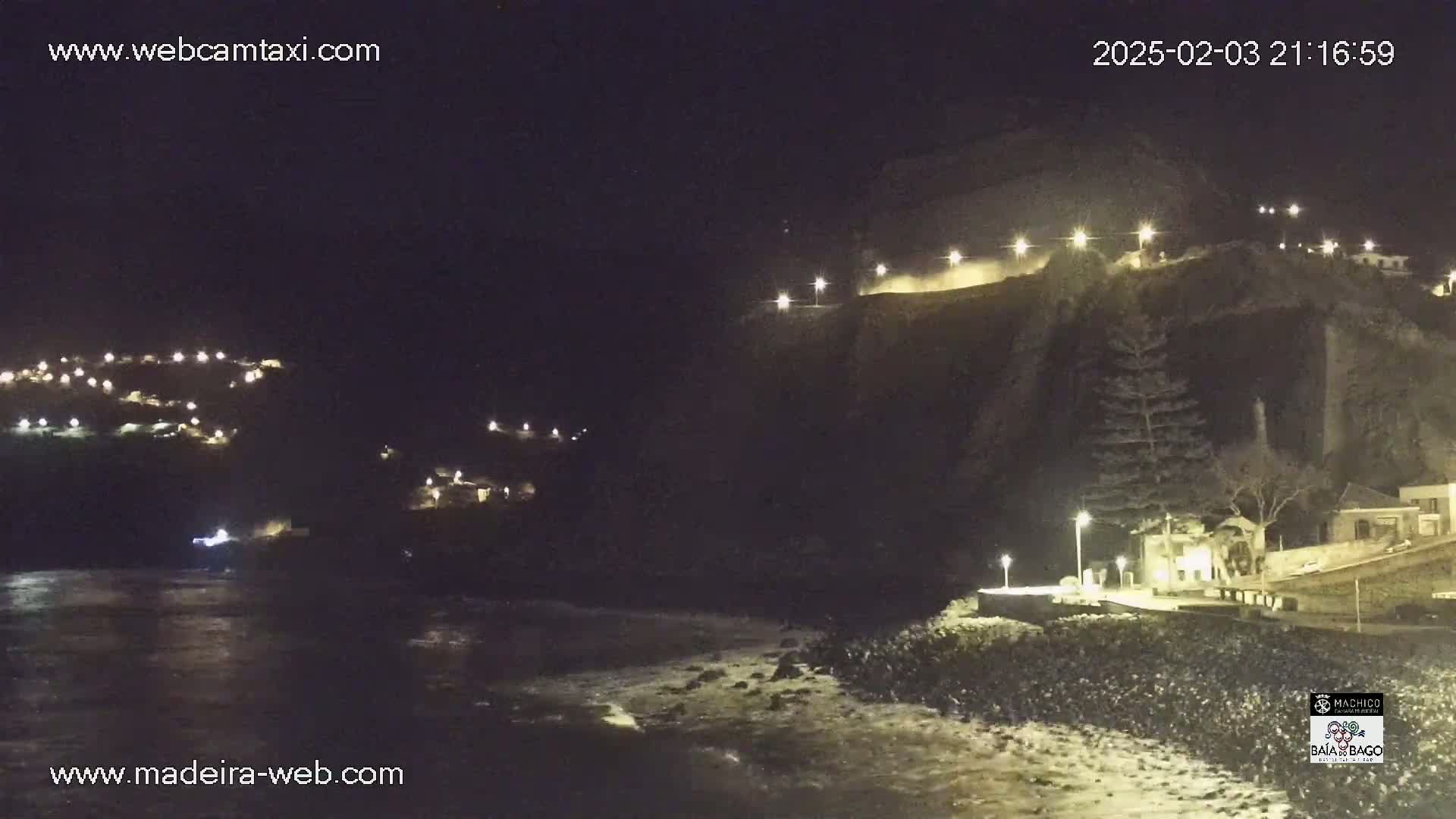 A nighttime coastal scene shows a dark cliff face illuminated by lights, a dark ocean with small waves, and small clusters of lights on the hillsides.