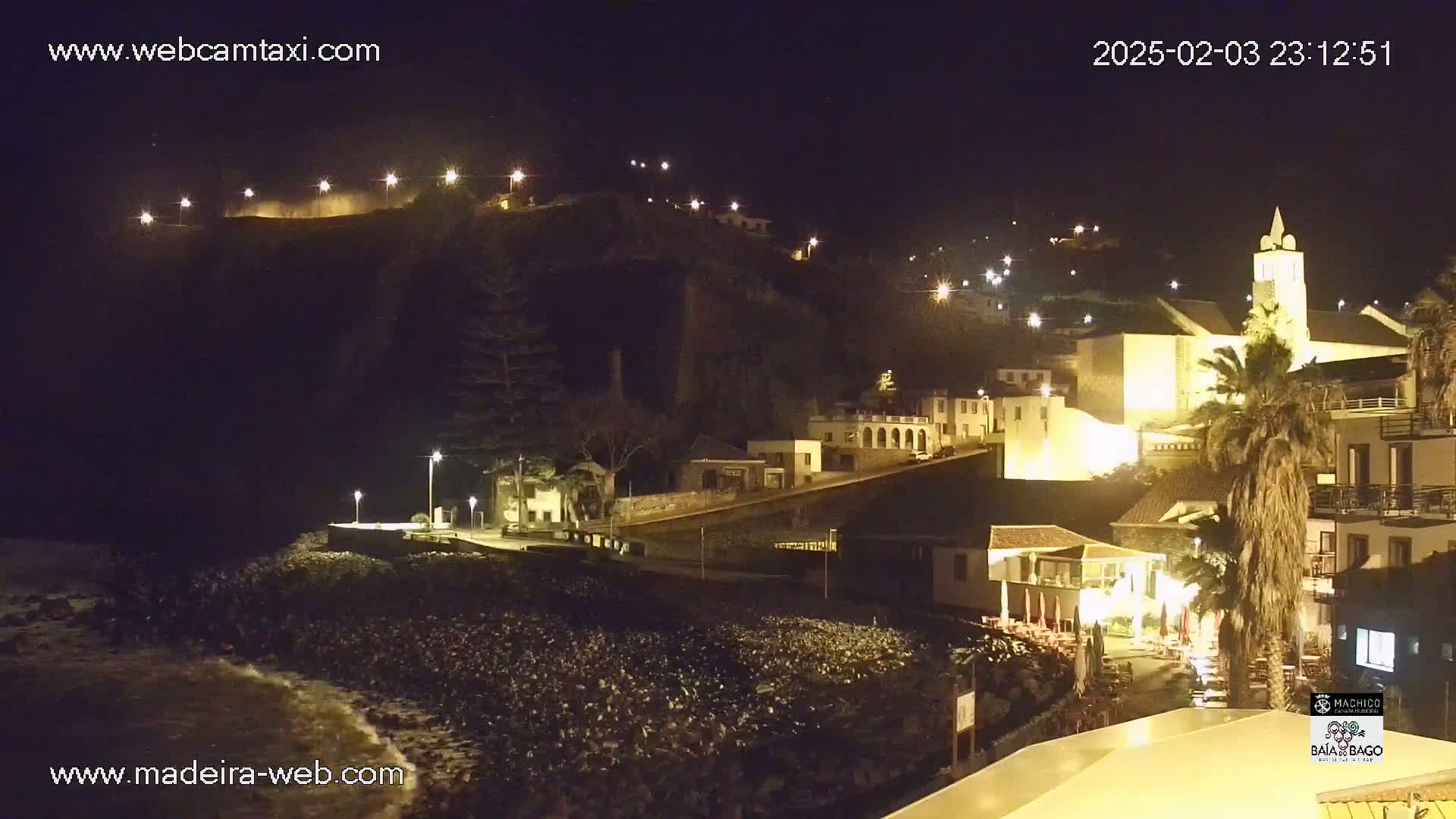 A nighttime coastal town with illuminated buildings, a church, and palm trees is shown, with gentle waves lapping at a dark, pebbled shore.
