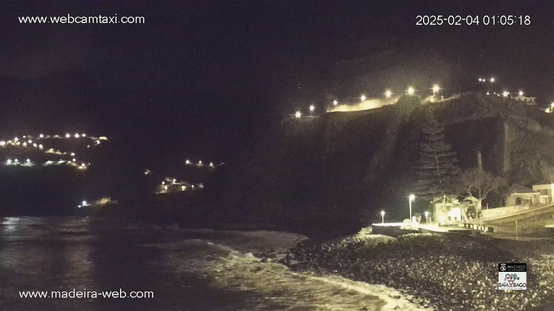 A nighttime coastal scene shows a dark cliff illuminated by lights, with gentle waves lapping a rocky shore.