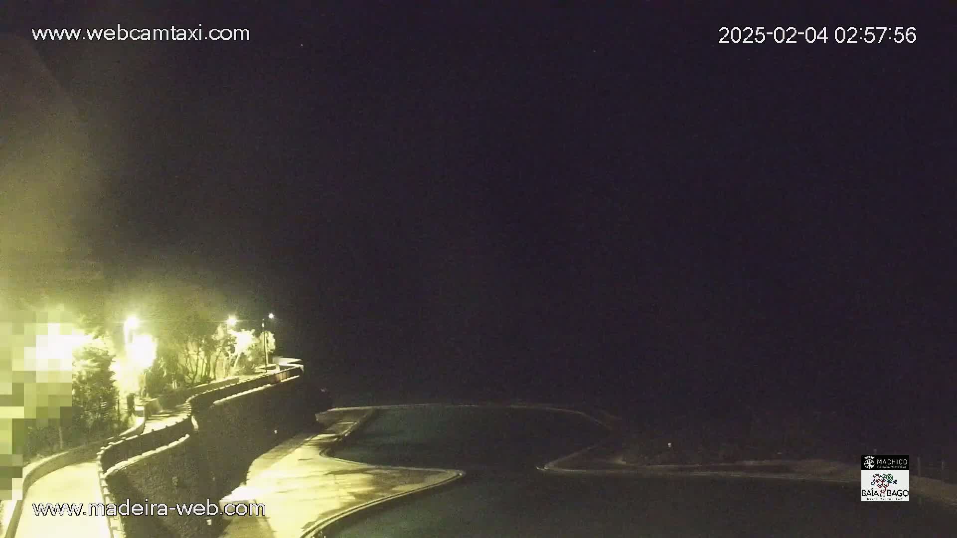 A nighttime view of a dark, empty swimming pool area next to a wall with lights illuminating vegetation and a path.