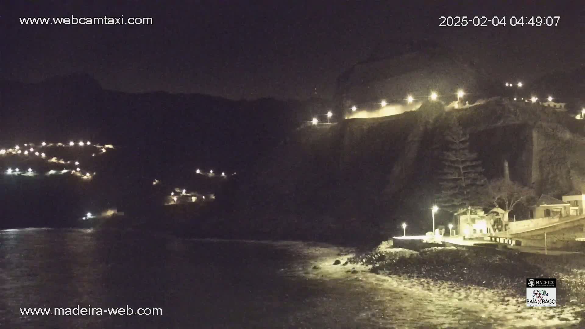 A nighttime coastal scene shows a dark cliff face with lights illuminating a road and buildings along its edge, overlooking a dark ocean with some small waves.