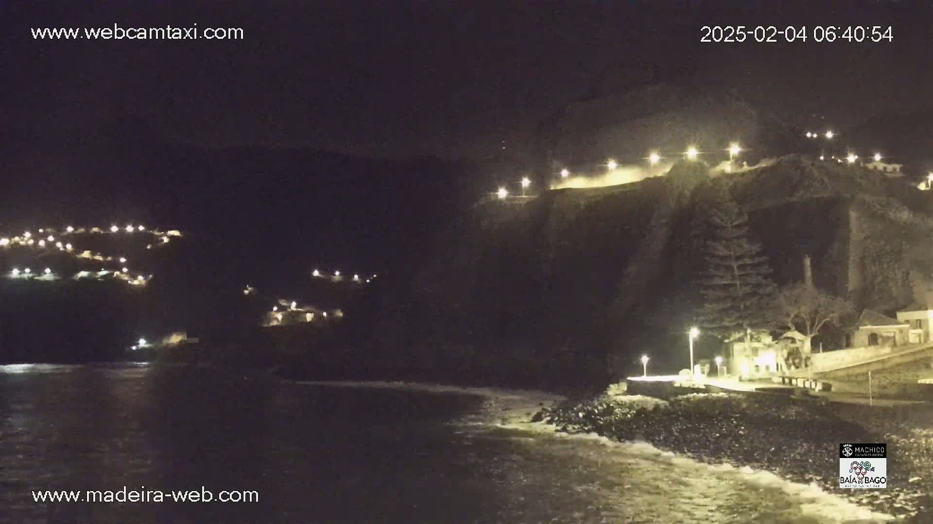 A nighttime coastal scene shows a dark cliffside with lights illuminating buildings and a roadway above a rocky shoreline with gentle waves.