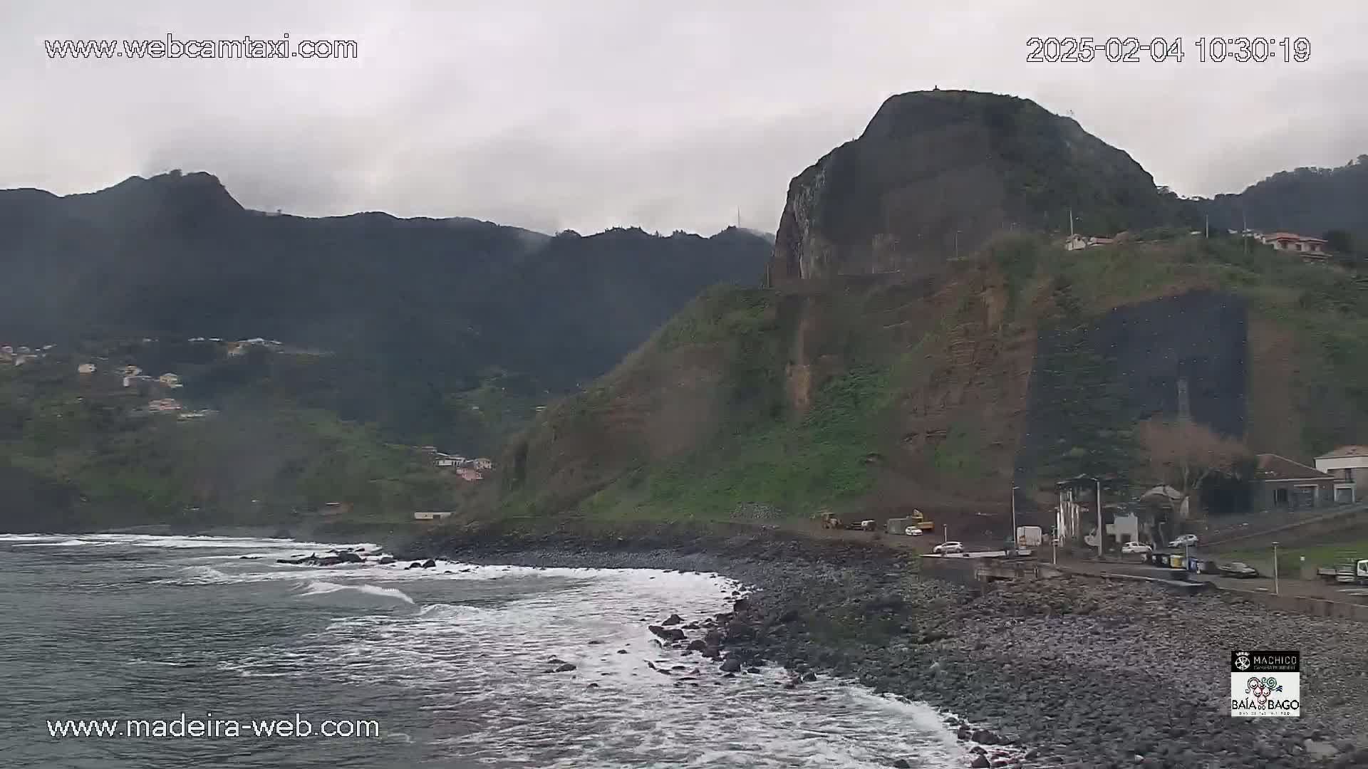 A rocky coastline with a dark, overcast sky meets dark green mountains, and some light waves lap against a pebble shore.