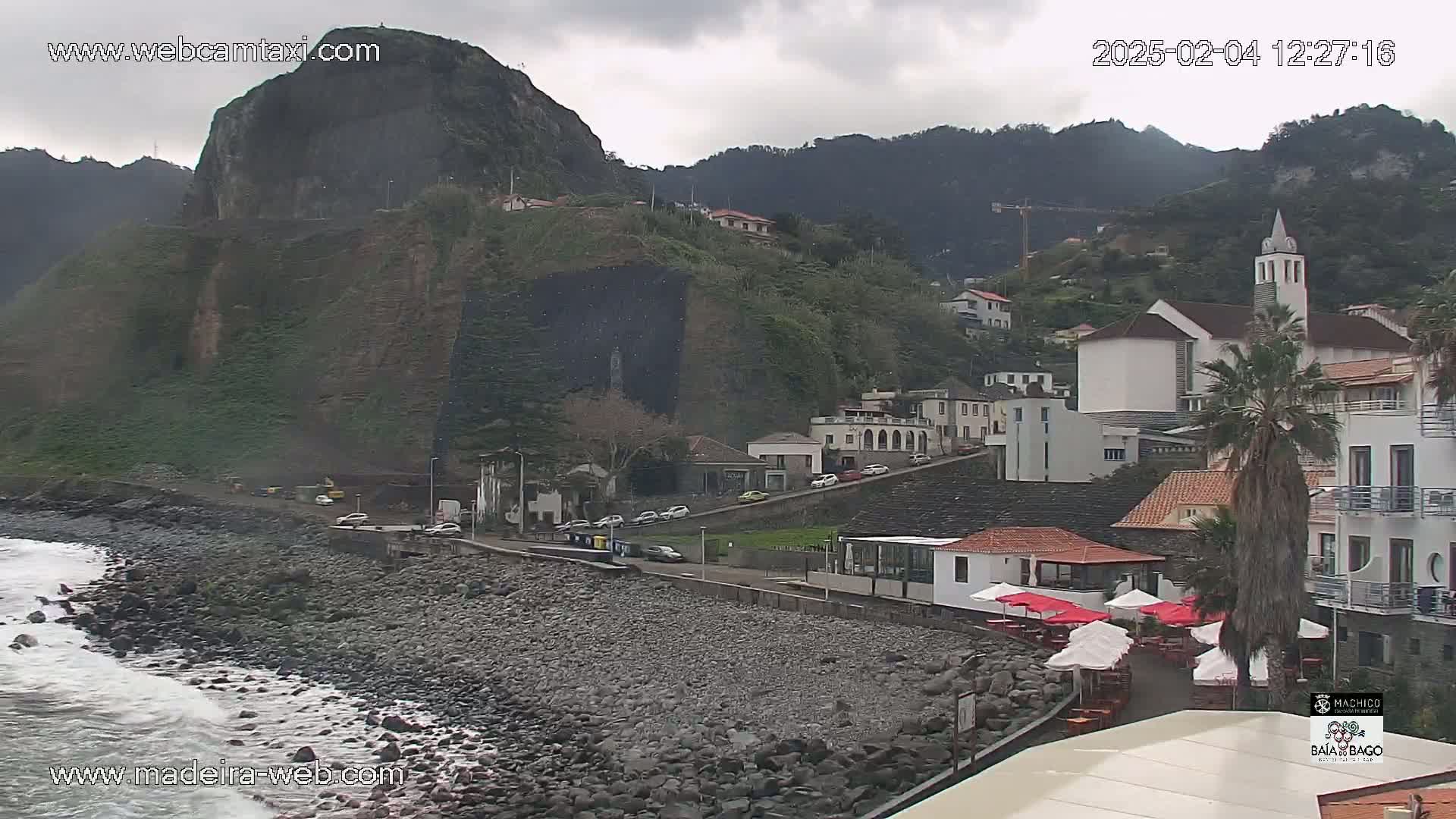 A rocky coastline with a village of white buildings nestled against a steep, dark cliff under an overcast sky.