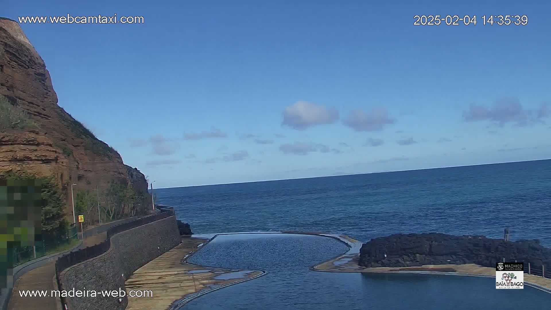 A rocky cliff overlooks a calm ocean with a man-made swimming pool situated between the cliff and the water under a mostly clear sky with a few scattered clouds.