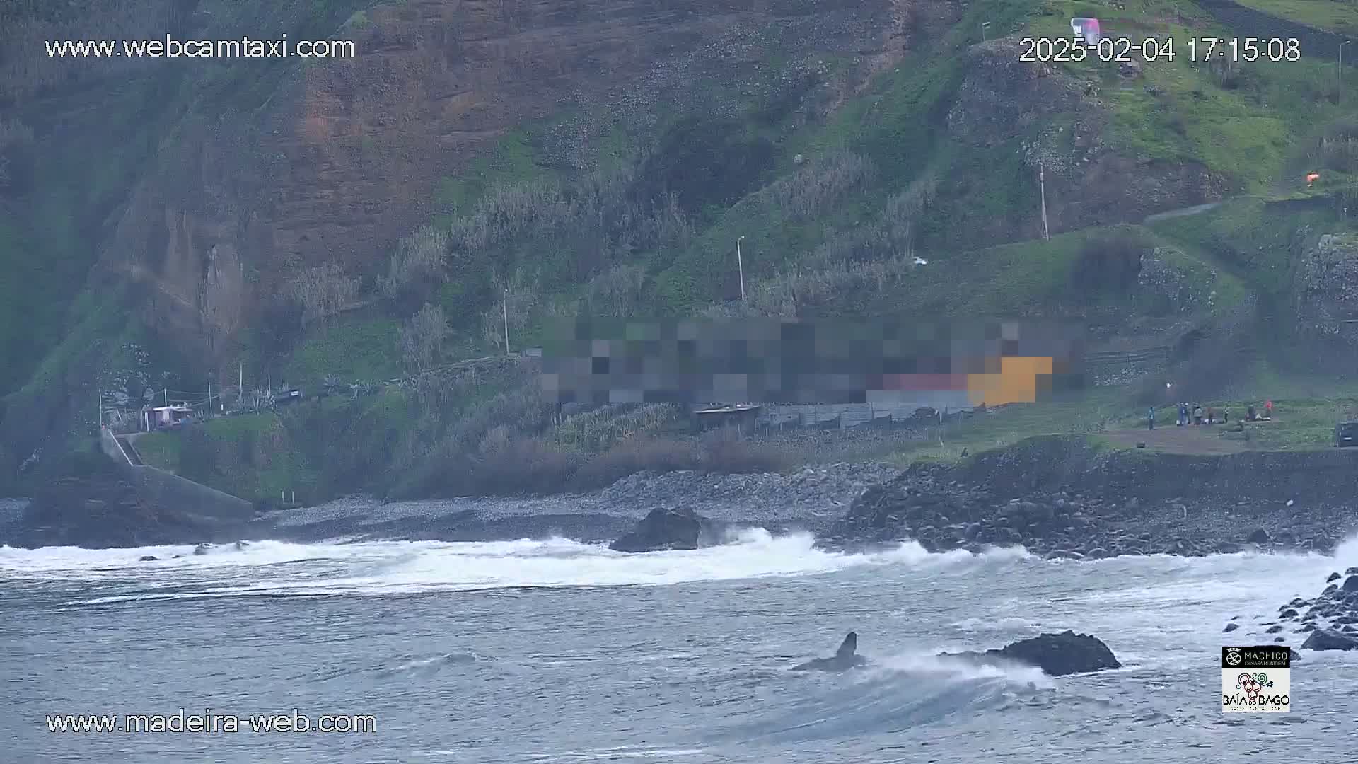 Ocean waves crash against a rocky shore near a hillside with some structures and a small group of people.
