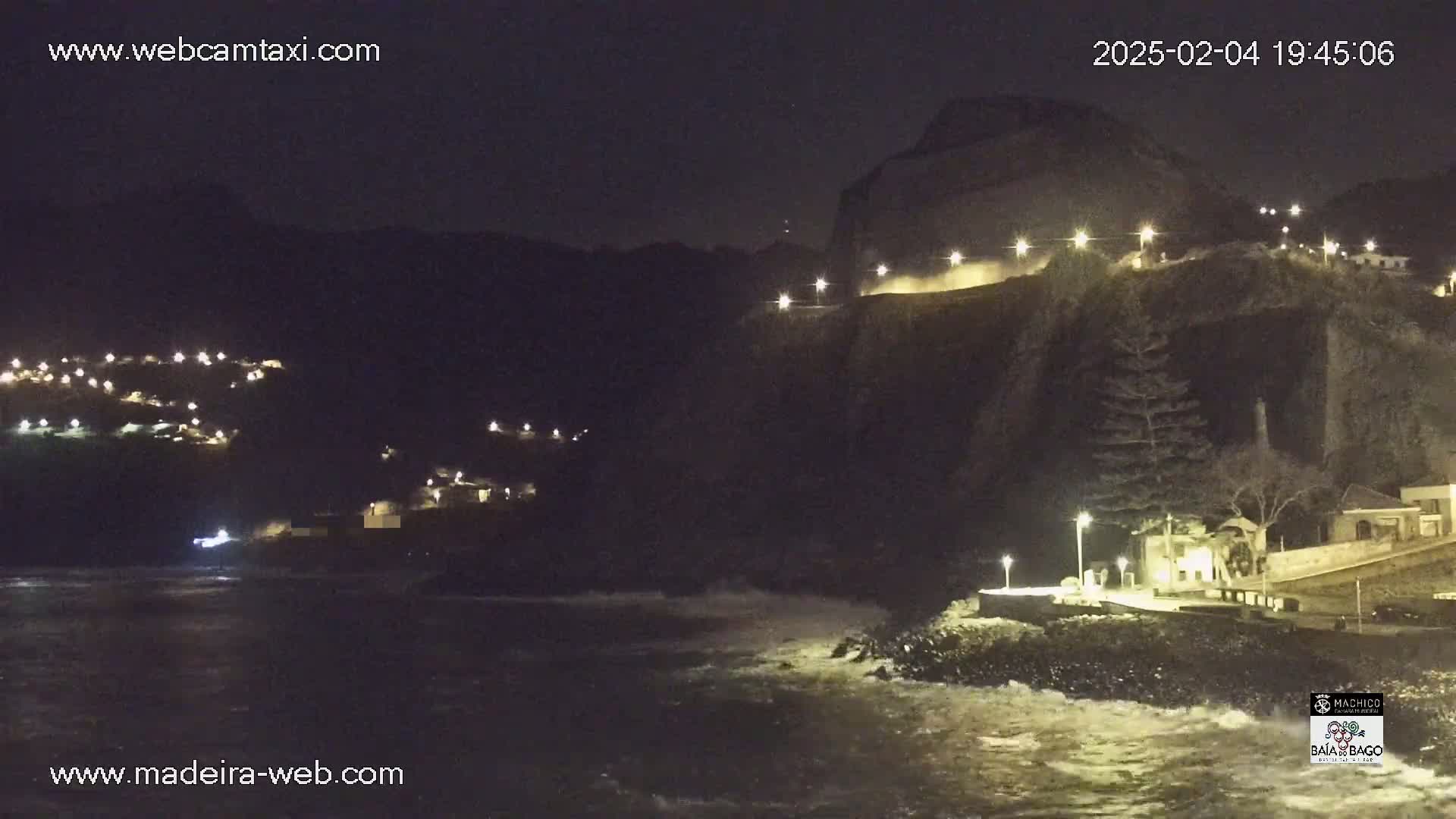 A nighttime coastal scene shows a dark cliff face with lights illuminating a roadway and buildings, along with dark water crashing against the shore below.