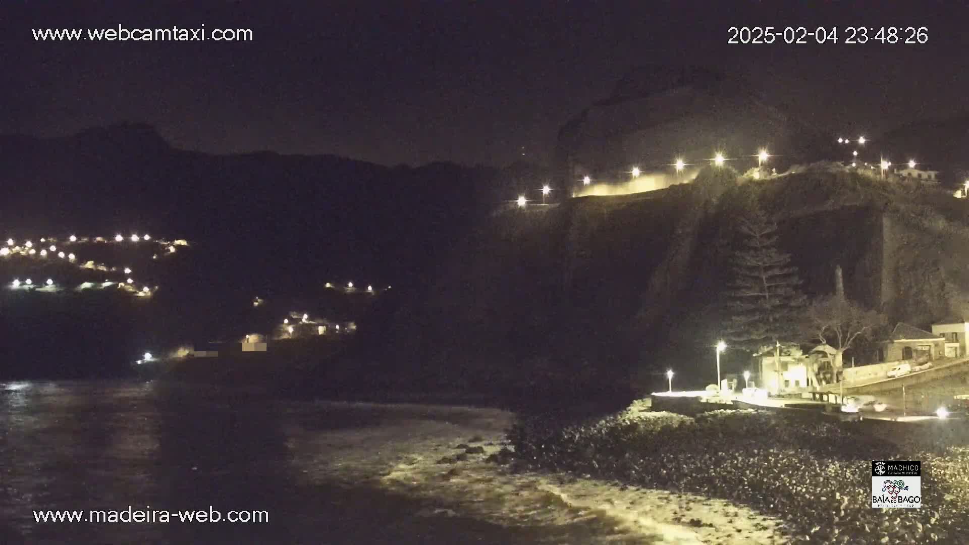 A nighttime coastal scene shows dark cliffs with illuminated buildings and roads above a rocky shoreline with waves gently lapping at the shore.