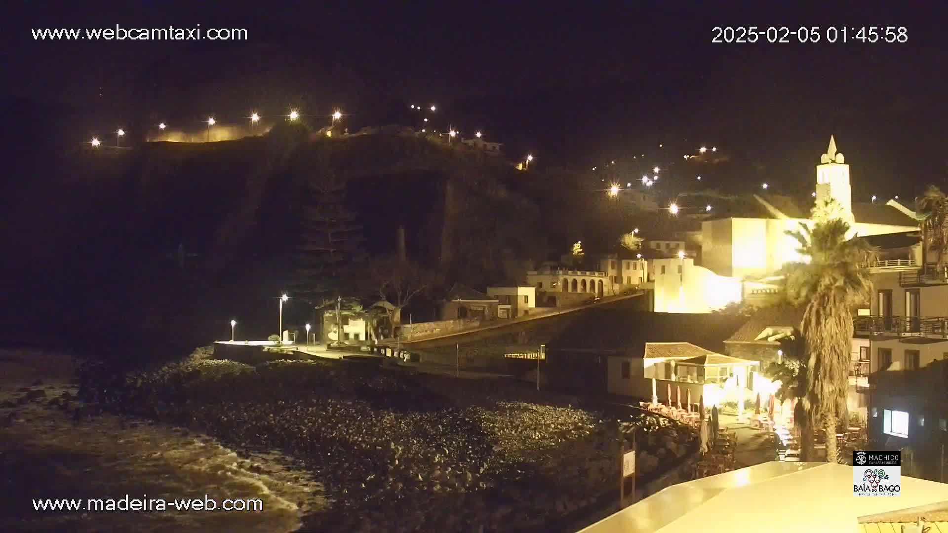 A nighttime view of a coastal town illuminated by streetlights, with dark water lapping at a rocky shore.