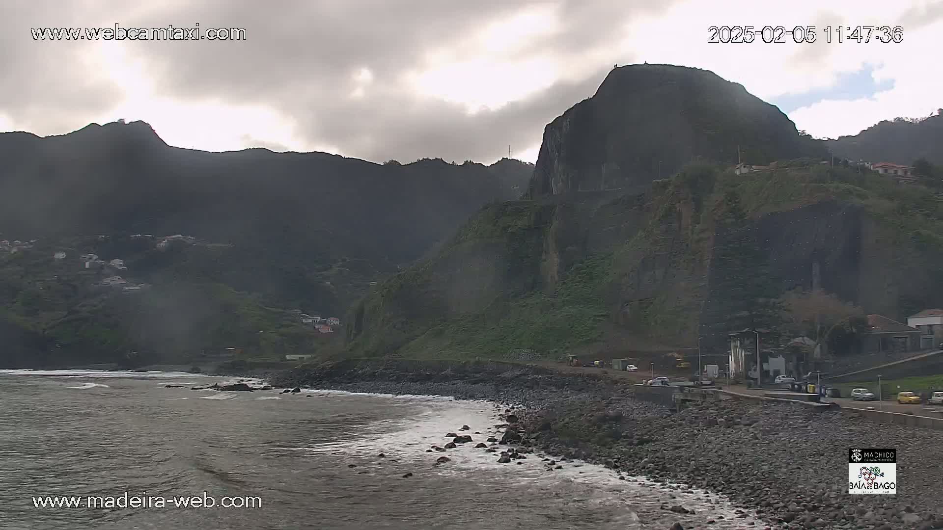 A rocky coastline with dark green hills and overcast skies meets a gray ocean with small waves.