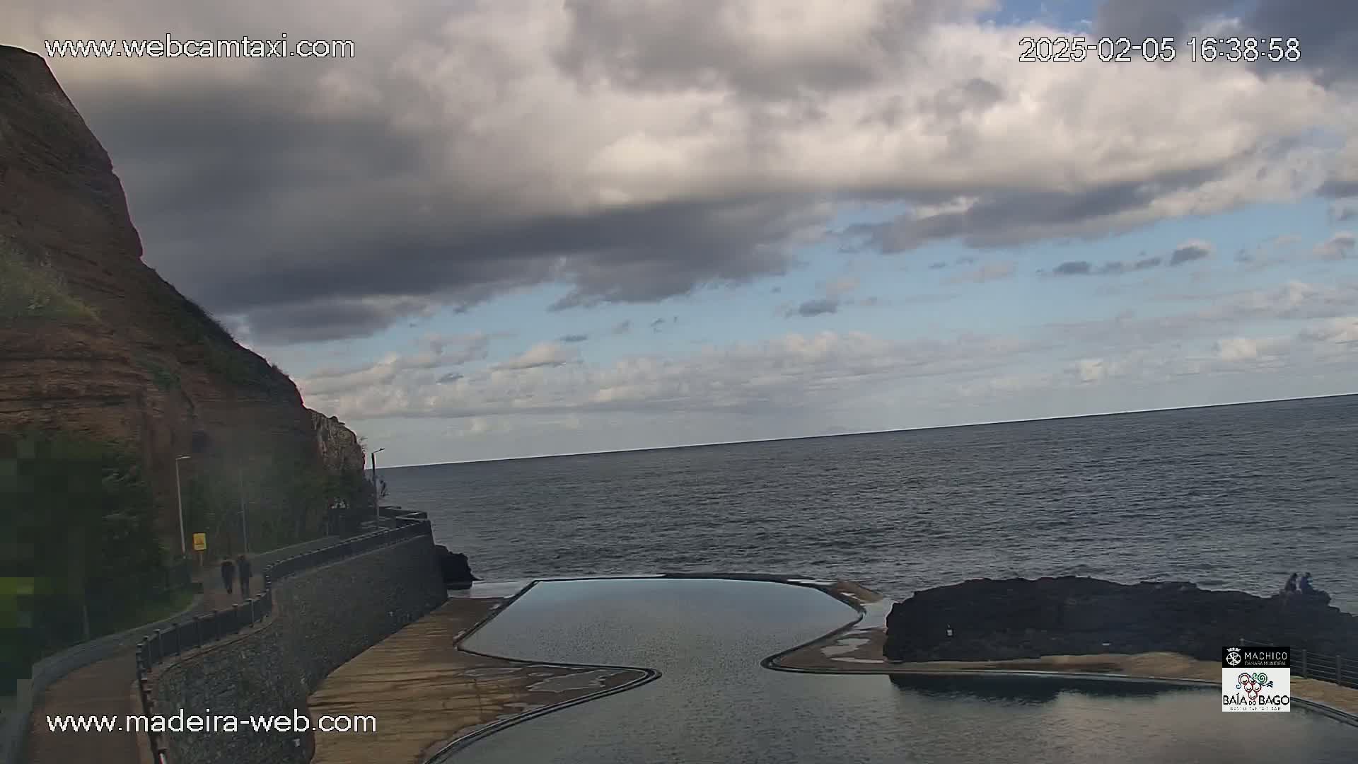 An outdoor scene shows a rocky cliffside overlooking a calm ocean under a mostly cloudy sky, with a naturally formed, kidney-shaped swimming pool nestled between the rocks and the water.