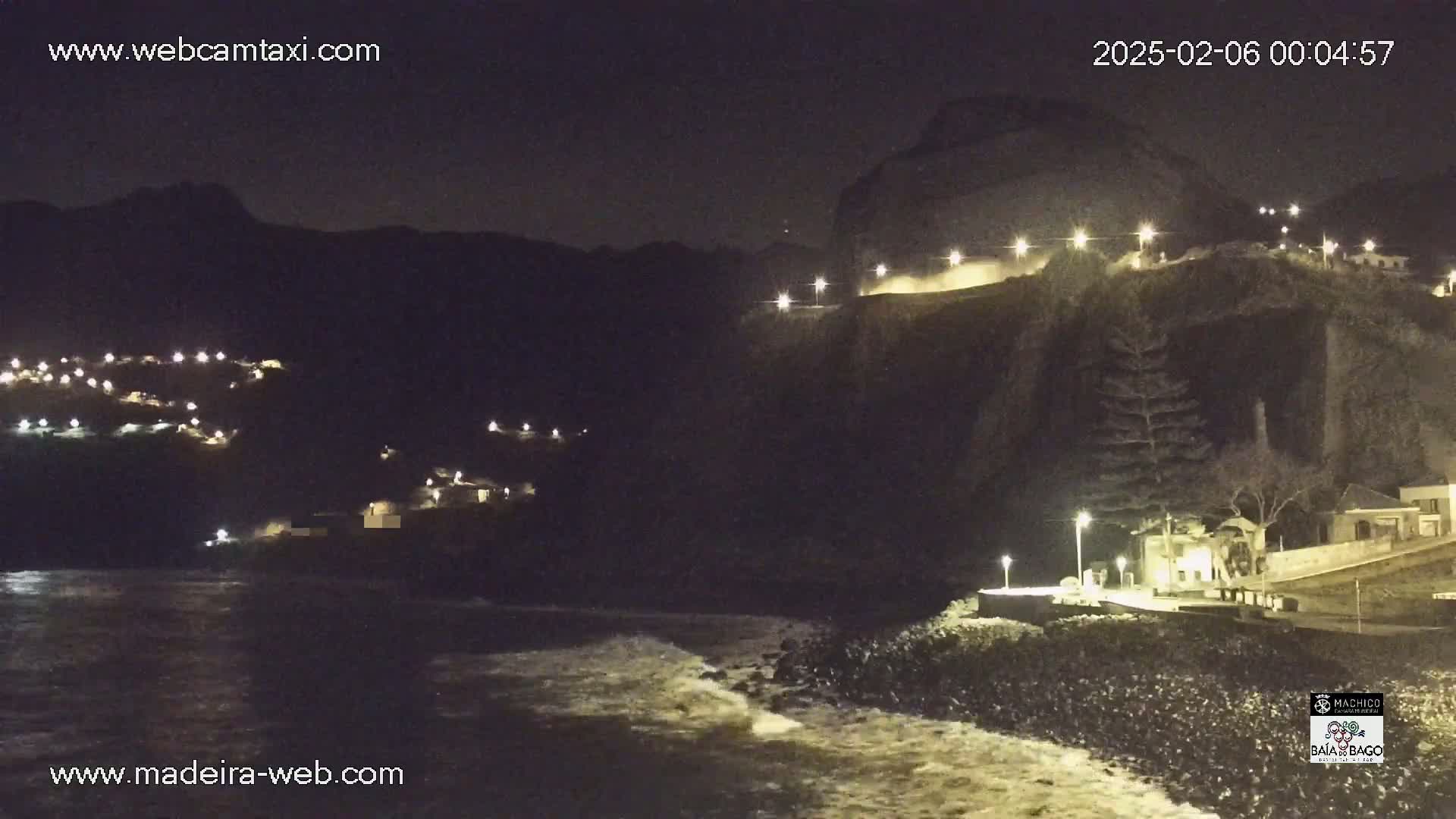 A nighttime coastal scene shows a dark, rocky cliff with lights illuminating buildings and a roadway, and small waves lapping at the shore.