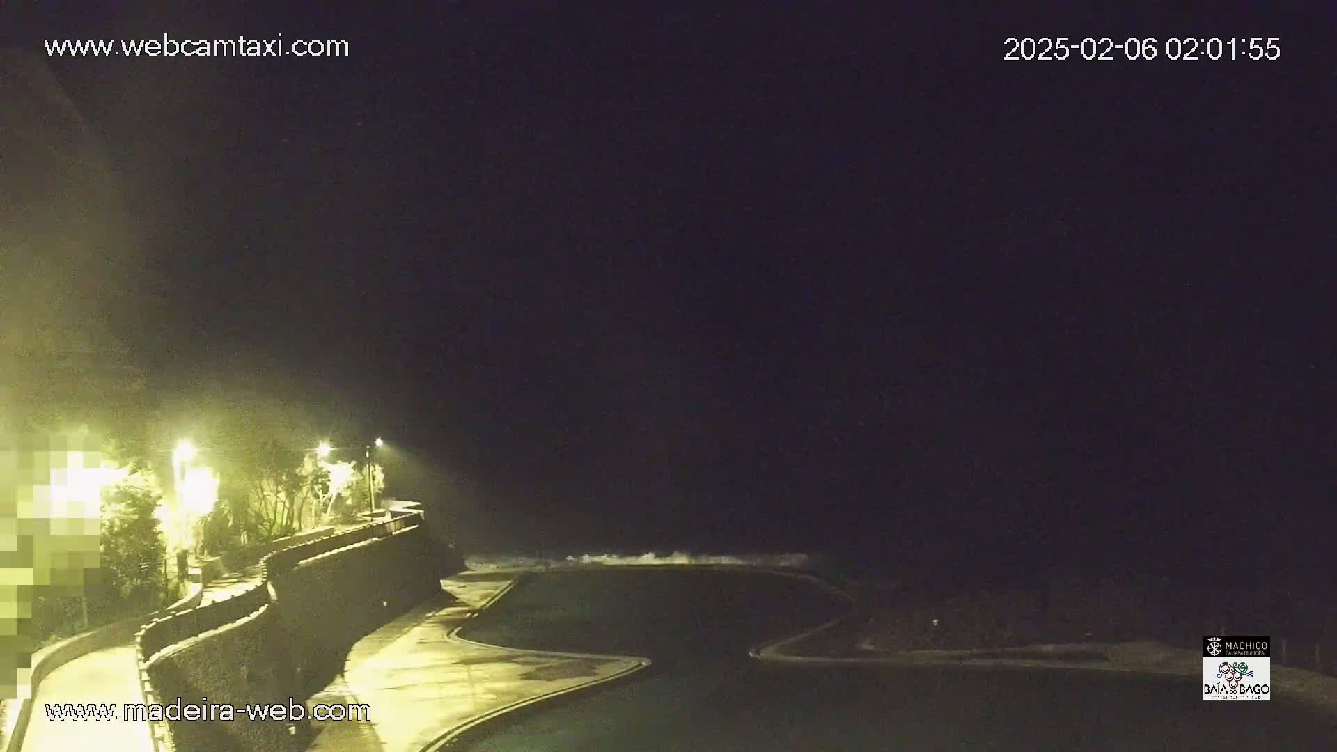 At night, a dark ocean with waves crashes near a paved walkway and an illuminated, empty swimming pool.