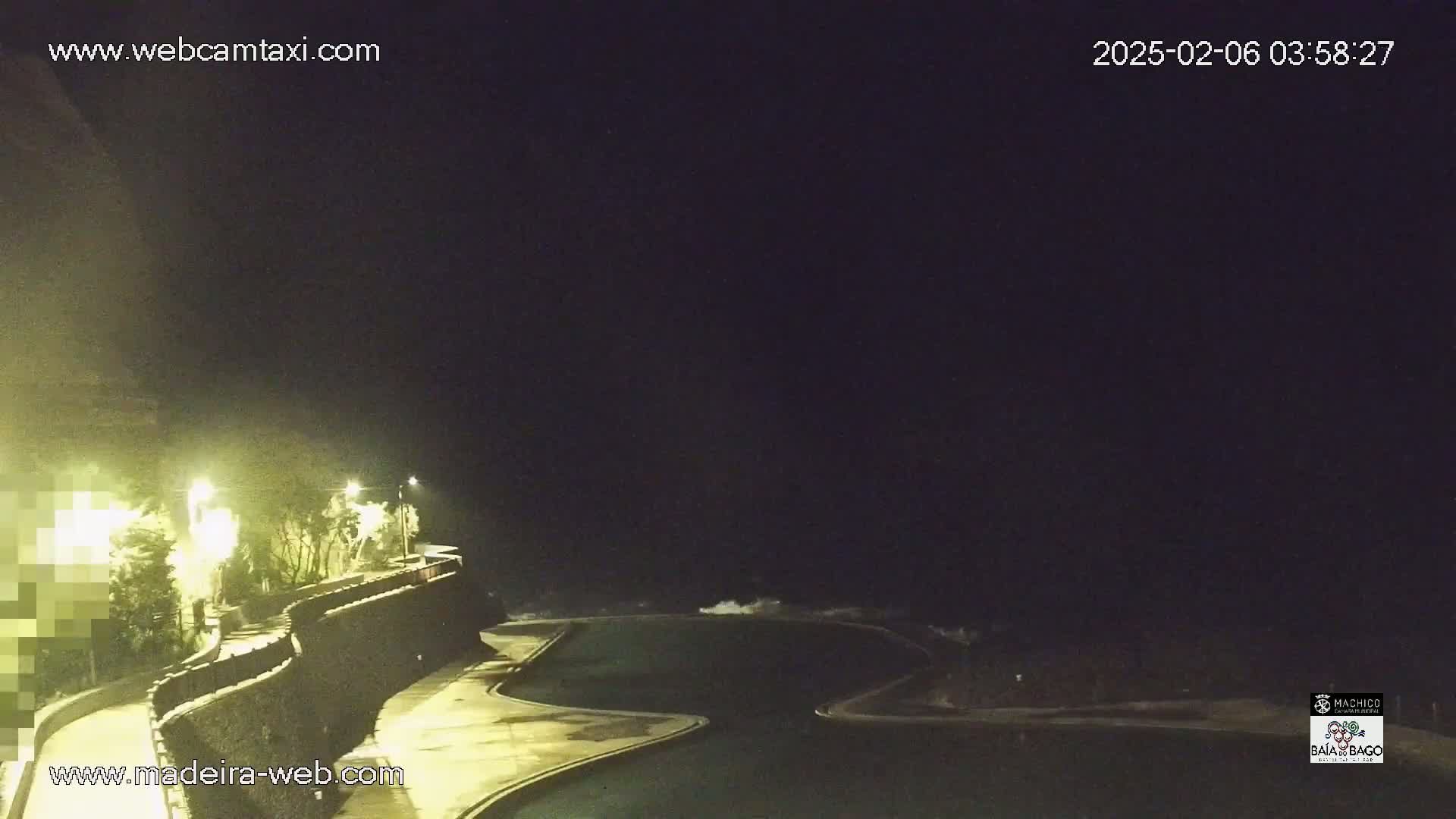 A nighttime view of a dark ocean with waves crashing near a lit, curved pool and walkway alongside a wall with vegetation.