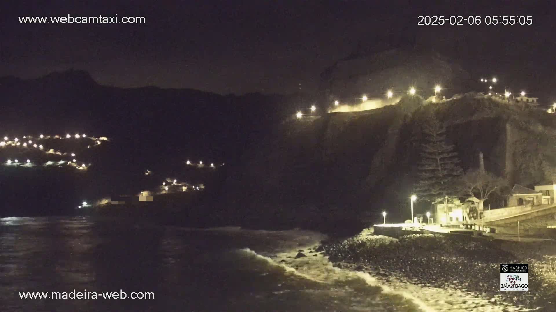 A nighttime coastal scene shows dark cliffs with illuminated areas, a dark ocean with small waves, and small clusters of lights on the hillsides.