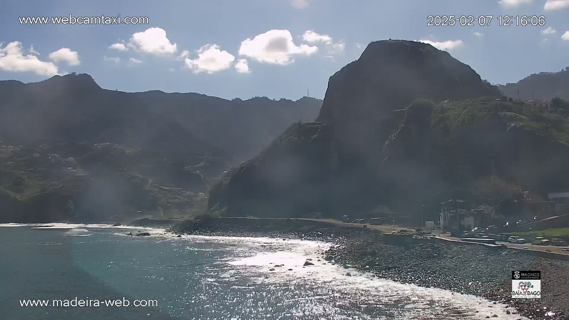 A rocky coastline with dark mountains in the background under a partly cloudy sky, with some small waves breaking on the shore.