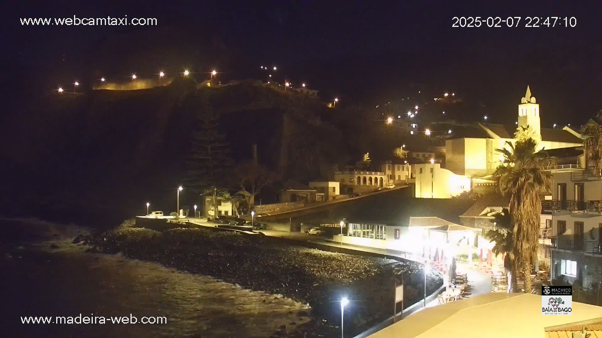 A nighttime coastal town scene with illuminated buildings, a church, palm trees, and gently lapping waves on a dark, rocky shore.
