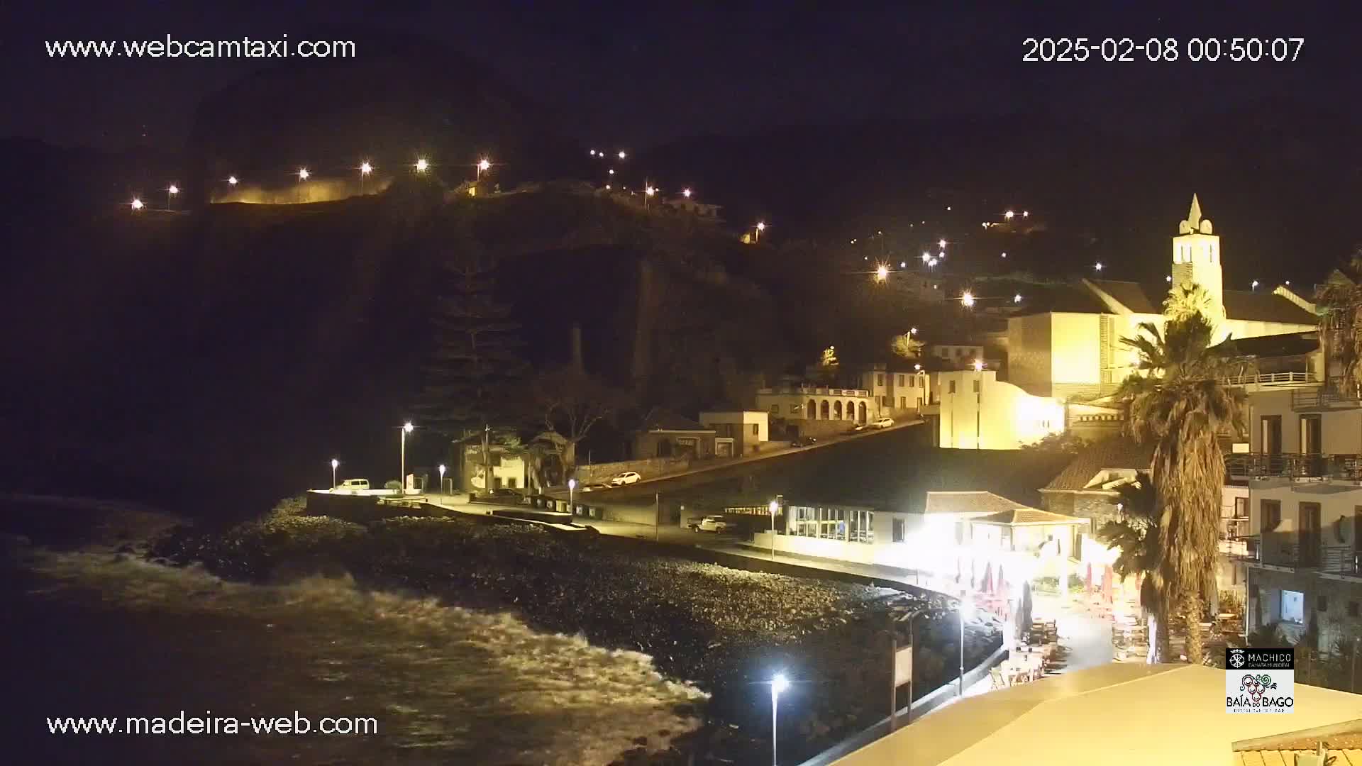 A nighttime coastal town with illuminated buildings and a church, overlooking a rocky shore with waves crashing against it.